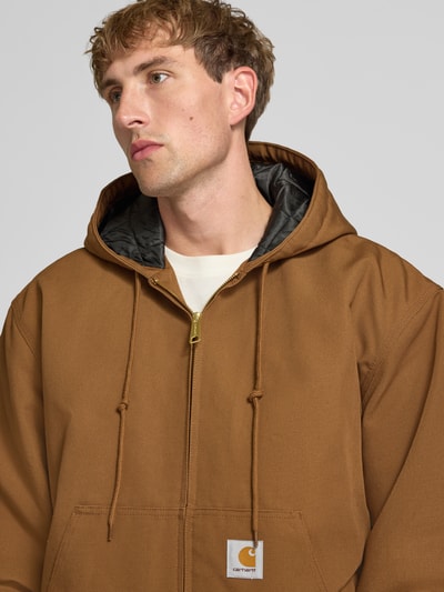Männermodel trägt Jacke mit Label-Patch und Känguru-Tasche von Carhartt Work In Progress in Braun, goldener Reißverschluss, Nahaufnahme Oberkörper. Reine Baumwolle, Kapuze mit Kordelzug, Regular Fit, gerippte Abschlüsse, Bildkontext integriert.