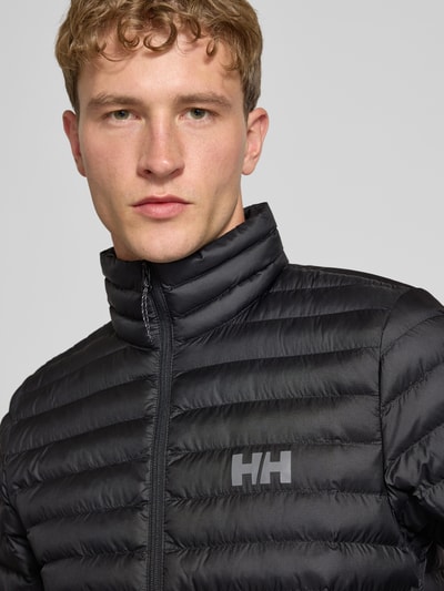 Männermodel trägt Steppjacke mit Label-Print von Helly Hansen, schwarze Farbe, Stehkragen, Nahaufnahme Oberkörper.