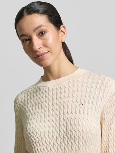 Frauenmodel trägt Slim Fit Pullover aus reiner Baumwolle von Tommy Hilfiger, Optik: Unifarben, Strickoptik, Zopfmuster, Logo-Detail, Rundhalsausschnitt, Langarm, Material: Strick, Farbe creme, Nahaufnahme Oberkörper.