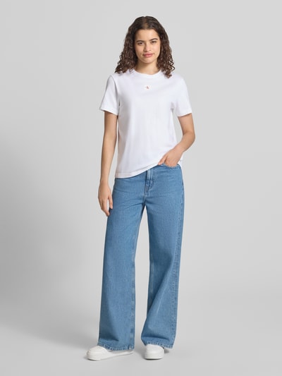 Frauenmodel trägt Regular Fit T-Shirt aus reiner Baumwolle von Calvin Klein Jeans in Weiß, kombiniert mit blauer Jeans, Ganzkörperansicht von vorne.