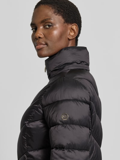 Vrouwenmodel draagt Steppmantel met Kapuze und Reißverschlusstaschen van MICHAEL Michael Kors, glanzend zwart, doorgestikte textuur, detailopname schouder en kraag, context: recht model, capuchon met tunnelkoord en cordstoppers, opstaande kraag, ritssluiting, zij-ritszakken, ruglengte maat S: 86 cm, schouderbreedte maat S: 43 cm, borstwijdte maat S: 50 cm, mouwlengte maat S: 62 cm.