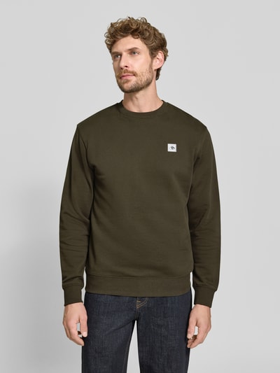 Męski model nosi Sweatshirt mit Label-Patch od Scotch & Soda z bawełny, luźny krój, okrągły dekolt, prążkowane wykończenia, długość tyłu 64 cm, długość rękawów 64 cm, widok całej sylwetki z przodu.