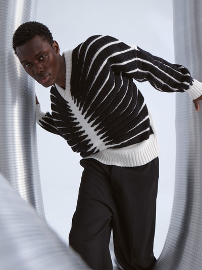 Männermodel trägt Pullover in Strick-Optik von GmbH mit Colour-Blocking-Design, V-Ausschnitt, lockerer Passform, Ganzkörperansicht von vorne.
