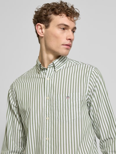 Męski model nosi Slim Fit Freizeithemd mit Brusttasche od Gant, białe i zielone paski, czysta bawełna, logo, krój slim fit, kieszonka na piersi, kołnierzyk button-down, długo rękaw, długość tyłu w rozmiarze M: 77 cm, szerokość ramion w rozmiarze M: 46 cm, długość rękawa w rozmiarze M: 65 cm, ujęcie z przodu, zbliżenie na górną część ciała.
