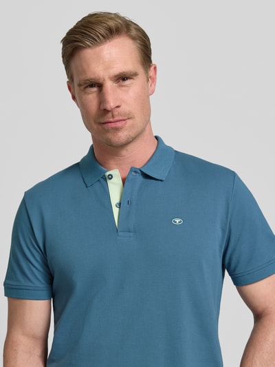Mężczyzna nosi Regular Fit Poloshirt aus reiner Baumwolle von Tom Tailor, efekt: jednolity kolor, krój regular fit, wyhaftowane logo, kołnierzyk polo, zapięcie na 2 guziki, krótki rękaw, wykończenie: z rozcięciami po bokach, wykończenie materiału: pika, materiał: czysta bawełna, kolor niebieski, gładka tekstura. Nahaufnahme Oberkörper.