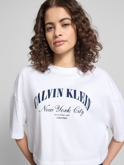 Frauenmodel trägt Cropped T-Shirt mit Logo-Print von Calvin Klein Jeans aus weißer Baumwolle mit Rundhalsausschnitt, Nahaufnahme Oberkörper.