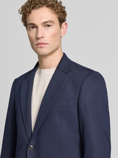 Męski model nosi marynarkę z mieszanki wiskozy z wpuszczanymi kieszeniami model 'JUSTIN' od Tiger Of Sweden, krój regular fit, kołnierz z połami, zapięcie na guzik, brustasza, długość tyłu w rozmiarze 50: 76 cm, szerokość ramion w rozmiarze 50: 36 cm, długość rękawów w rozmiarze 50: 66 cm, kolor granatowy, gładka tekstura. Zbliżenie na górną część tułowia.