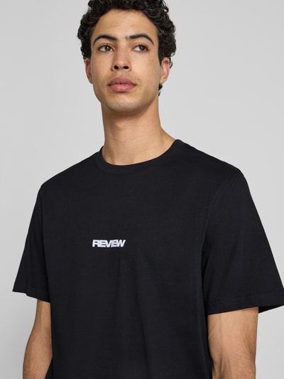 Męski model nosi T-Shirt mit Logo und Rundhalsausschnitt von REVIEW, czarna bawełna, krój relaxed fit, długość tyłu 68 cm, Nahaufnahme Oberkörper.