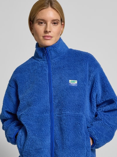 Frauenmodel trägt Jacke aus Teddyfell Modell 'HOKTOWN' von American Vintage in Blau mit Stehkragen, Reißverschluss, Label-Detail, Nahaufnahme Oberkörper.