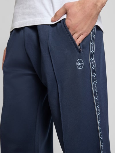 Männermodel trägt dunkelblaue Trainingshose mit Biese, aufgesetztem Band und Logo-Bestickung von REVIEW, Nahaufnahme Oberschenkel.