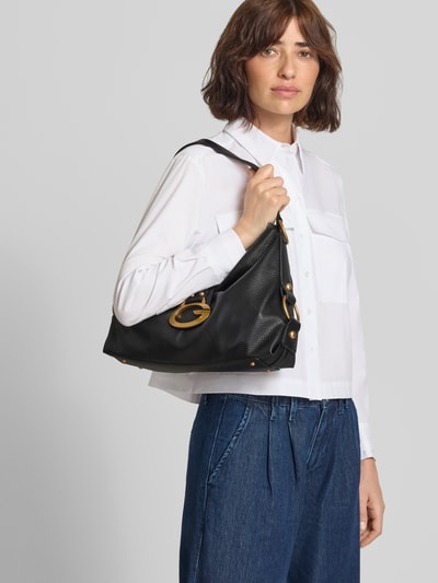 Frauenmodel trägt Handtasche mit Label-Applikation Modell 'CAMDEN' von GUESS in Schwarz mit Reißverschluss und Magnetverschluss, Nahaufnahme Oberkörper.