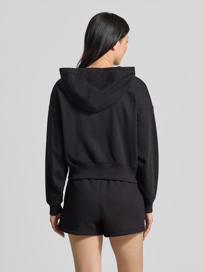 Frauenmodel trägt schwarze Regular Fit Sweatjacke mit Logo-Stitching Modell 'Heritage' von GAP aus Baumwoll-Mix, Kapuze mit Tunnelzug, geteilte Känguru-Tasche und elastische Abschlüsse, Ganzkörperansicht Rückansicht.