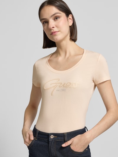 Kobiece model prezentuje damski t-shirt z mieszanki bawełny i elastanu, dopasowany krój, wyhaftowane logo, model 'SCRIPT' od Guess, dekolt w łódkę, długość tyłu w rozmiarze S: 61 cm, jasny kolor, gładka tekstura, pełny przód.