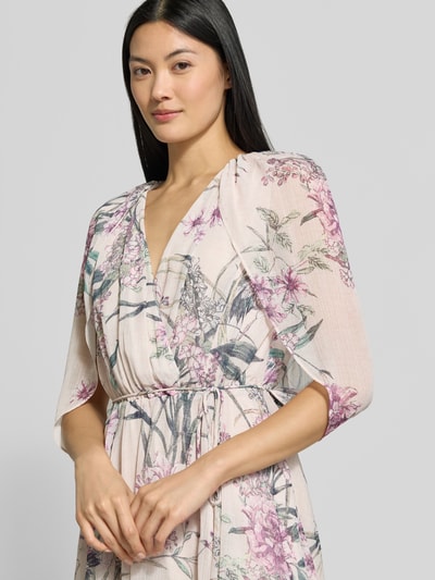 Vrouwenmodel draagt avondjurk met V-hals model 'Jodie' van Forever New, chiffon, getailleerde pasvorm, wikkellook, halflange vleermuismouwen, maxilengte, smalle tailleband, gevoerde onderrok, ruglengte maat 36: 135 cm, borstwijdte maat 36: 30 cm, bloemmotief in roze en groen, nahaufnahme bovenlichaam.