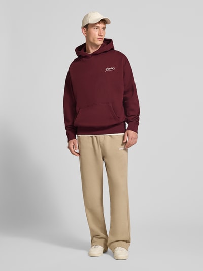 Männermodel trägt Regular Fit Hoodie aus Baumwoll-Mix von PEGADOR in Dunkelrot mit Kängurutasche und Strass-Logo, Ganzkörperansicht von vorne.