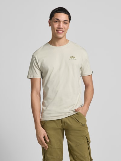 Männermodel trägt T-Shirt aus Baumwolle von Alpha Industries in Beige mit Label-Print-Details, Gerader-Schnitt, Rundhalsausschnitt, Ganzkörperansicht von vorne.