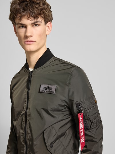 Męski model nosi Regular Fit Bomberjacke mit Logo-Patch od Alpha Industries w kolorze oliwkowym, z naszywką logo, kieszeniami, zamkiem i kieszenią na rękawie, materiał o gładkiej fakturze, krój regular fit, długość tyłu 65 cm, szerokość ramion 45 cm, szerokość w rozmiarze M: 54 cm, długość rękawa 69 cm, nahaufnahme górnej części ciała.