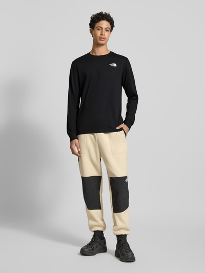 Męski model nosi Regular Fit Longsleeve mit Logo-Print od The North Face, czarny bawełniany longsleeve, krój regular fit, logo na piersi, okrągły dekolt, długie rękawy, elastyczne mankiety, długość tyłu w rozmiarze M: 69 cm, widok całej sylwetki z przodu.