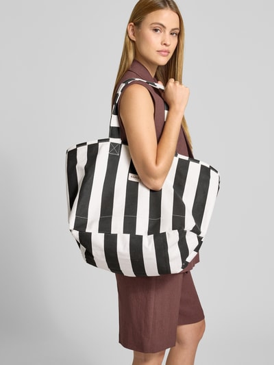 Kobiece model prezentuje torbę shopper w wzór na całej powierzchni, model 'JUNO' od Barts, czarno-białe pasy, czysta bawełna, uchwyt, szerokość w rozmiarze 1: 35 cm, długość w rozmiarze 1: 37 cm, głębokość w rozmiarze 1: 35 cm, długość paska na ramię w rozmiarze 1: 57 cm, ujęcie całej sylwetki z przodu.