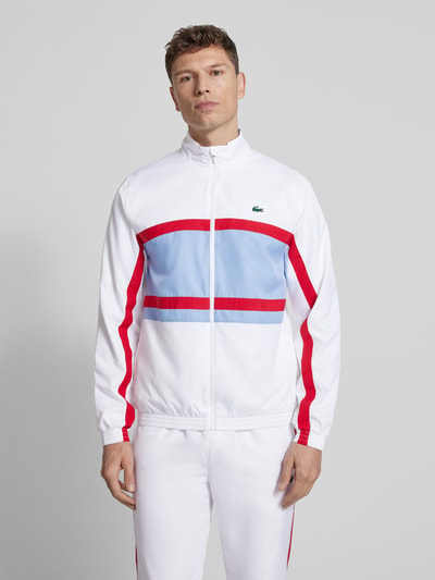 Männermodel trägt Regular Fit Trainingsanzug mit Kontraststreifen von Lacoste Sport in Weiß mit roten und blauen Details, Ganzkörperansicht von vorne.
