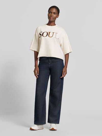 Frauenmodel trägt Cropped Boxy Fit Sweatshirt mit Label-Stitching von Smith and Soul, beige mit braunem Schriftzug, Damen Sweatshirt aus Baumwoll-Elasthan-Mix, Rundhalsausschnitt, 1/2-Arm, kombiniert mit dunkler Jeans und weißen Sneakern, Ganzkörperansicht von vorne.