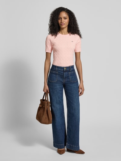 Frauenmodel trägt Strickshirt mit Zopfmuster und Label-Stitching von Gant aus Baumwoll-Mix mit Stretch-Anteil, Slim-Fit, Rundhalsausschnitt in Rosa, kombiniert mit blauer Jeans, Ganzkörperansicht von vorne.
