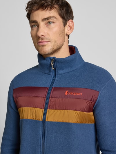 Männermodel trägt Jacke mit Stehkragen Modell 'Teca' von Cotopaxi in Blau mit farbigen Streifen, Label-Print und Reißverschluss, Nahaufnahme Oberkörper. Regular Fit, Eingrifftaschen, Rückenlänge 68 cm, Schulterbreite 43 cm, Brustweite 42 cm, Ärmellänge 63 cm.
