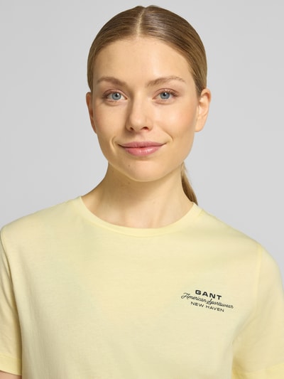 Kobieta nosi T-Shirt mit Label-Print und Rundhalsausschnitt od Gant z bawełny, krój regular fit, nadruk z logo, okrągły dekolt, długość tyłu w rozmiarze S 57 cm, jasny kolor, gładka tekstura, Nahaufnahme górnej części ciała.