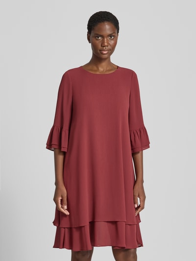 Frauenmodel trägt Relaxed Fit Kleid in Knielänge von Christian Berg Cocktail in Dunkelrot mit Rüschenbesatz, Halbarm, Ganzkörperansicht von vorne.