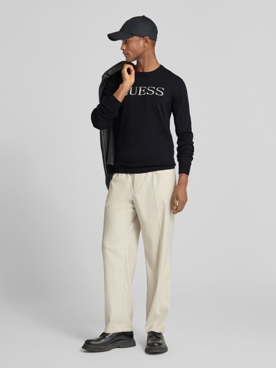 Männermodel trägt Regular Fit Pullover in Strick-Optik Modell 'Kane' von Guess aus reiner Baumwolle, Logo-Stitching, Rundhalsausschnitt, Ganzkörperansicht von vorne.