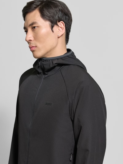 Männermodel trägt Regular Fit Funktionsjacke aus wasserdichtem Material Modell 'L_COMMUTER' von BOSS Green in Schwarz, Optik: Unifarben, Label-Print, reflektierende Details, seitliche Reißverschlusstasche, Kapuze und langarm, Nahaufnahme Oberkörper.