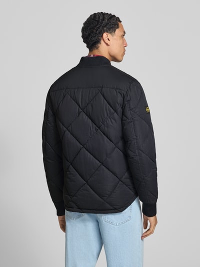 Männermodel trägt Regular Fit Steppjacke mit opstaande kraag van Superdry in zwarte Stiksels-look met steekzakken, elastische mouwboorden en ritssluiting, ruglengte maat M 69 cm, schouderbreedte 46 cm, borstwijdte 58 cm, mouwlengte 59 cm, geheel van achteren