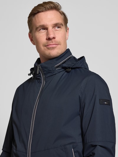 Mannenmodel draagt 'Regular Fit Jacke mit Stehkragen' van bugatti in donkerblauw, met logodetail, capuchon, tweewegritssluiting, steekzakken, elastische mouwboorden, regular fit; context: heren jack, ruglengte 68 cm, schouderbreedte 42 cm, borstwijdte 50 cm, mouwlengte 65 cm. Nahaufnahme bovenlichaam.