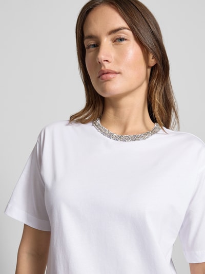 Modelka kobieca nosi białe T-Shirt z bawełny JOOP!, luźny krój, okrągły ściągacz i detal z logo, model 'Thei', zbliżenie górnej części ciała.