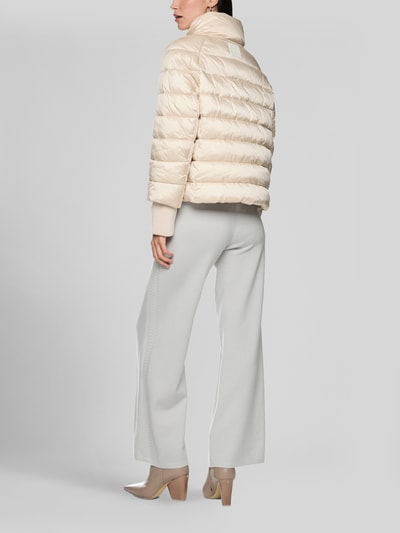 Frauenmodel trägt Steppjacke mit Stehkragen und Label-Detail von Marc Cain in Beige, strukturierte Oberfläche, Ganzkörperansicht Rückansicht. Rückenlänge 56 cm, Schulterbreite 41 cm, Brustweite 55 cm, Ärmellänge 73 cm, reguläre Passform, Eingrifftaschen.