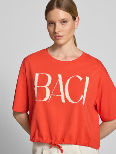 Frauenmodel trägt Boxy Fit T-Shirt mit Perlen und Tunnelzug von Oui, Damen T-Shirt aus Baumwoll-Elasthan-Mix, Rundhalsausschnitt, Rückenlänge 51 cm bei Größe 36, leuchtendes Orange, weiße BACI-Aufschrift, glatte Textur, Nahaufnahme Oberkörper.