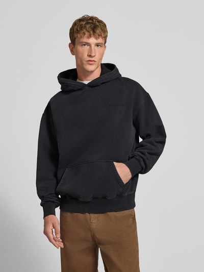 Männermodel trägt Oversized Hoodie aus Baumwoll-Mix von PEGADOR in Schwarz mit überschnittenen Schultern und Logo-Stickerei, Ganzkörperansicht von vorne. Rückenlänge 68 cm, Ärmellänge 70 cm, gerippte Bündchen, lockere Passform.