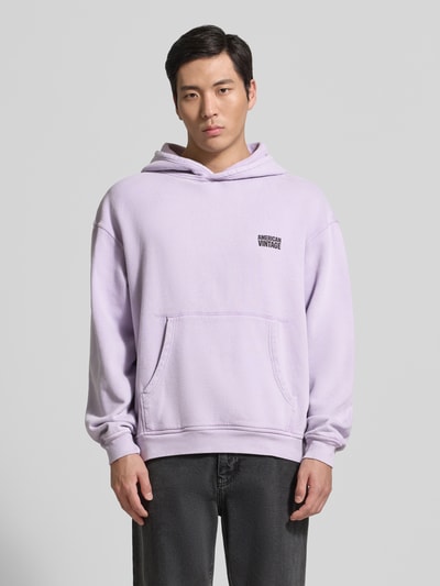 Męski model nosi bluzę z kapturem z logo model 'PLIZZY' od American Vintage z bawełny, krój relaxed fit, kieszeń kangurka, lawendowy kolor, długość tyłu 66 cm, długość rękawów 60 cm, widok całej sylwetki z przodu.