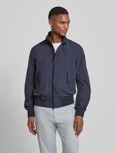 Męski model nosi Jacke mit Pattentaschen Modell 'Alwin' od Blauer Usa, krój regular fit, granatowy kolor, stehkraag, naszywki z logo, kieszenie z patkami zapinane na guziki, dwukierunkowy zamek, prążkowane wykończenia, szerokość w rozmiarze M: 54 cm, długość tyłu: 66 cm, szerokość ramion: 47 cm, długość rękawa: 67 cm; całkowity widok z przodu.