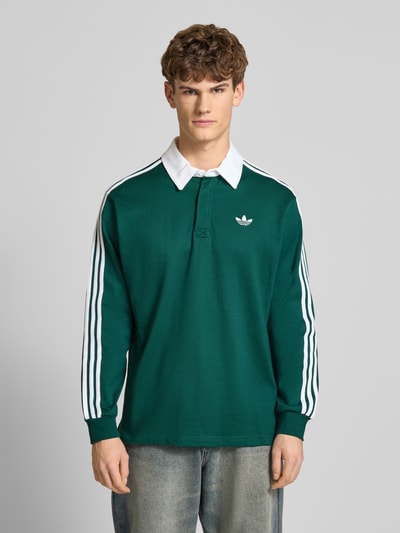 Männermodel trägt Poloshirt mit kurzer Knopfleiste von adidas Originals in Dunkelgrün mit weißen Streifen, reine Baumwolle, Ganzkörperansicht von vorne.