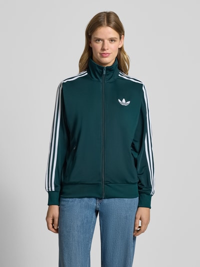Frauenmodel trägt Bluza rozpinana z stójką od adidas Originals, luźny krój, zamek błyskawiczny, wyhaftowane logo, kieszenie zapinane na zamek błyskawiczny, prążkowane wykończenia, kolor ciemnozielony z białymi akcentami, gładka tekstura, Ganzkörperansicht von vorne.
