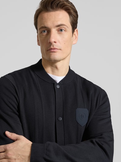 Męski model nosi Regular Fit Strickjacke aus Viskose-Mix Modell 'KN_BB BC LUX FZ' od BOSS Green, czarny, prążkowana dzianina, naszywka z logo, zatrzaski, college’owy kołnierz, długie rękawy, materiał: mieszanka wiskozy. Zbliżenie na górną część ciała.