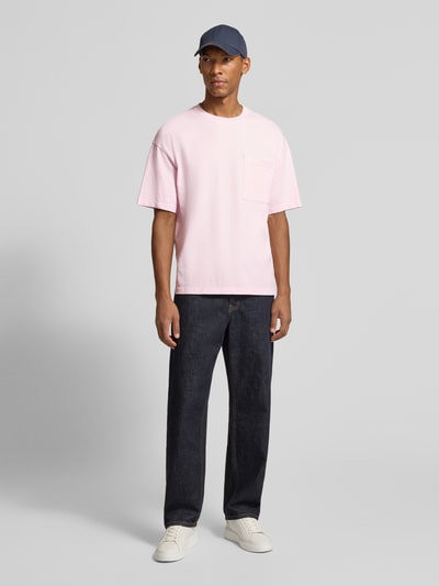 Männermodel trägt T-Shirt aus reiner Baumwolle mit Brusttasche Modell 'AFOMA' von American Vintage in Hellrosa, dunkle Hose, Ganzkörperansicht von vorne, Relaxed Fit, Rundhalsausschnitt, Halbarm, überschnittene Schultern, Rückenlänge bei Größe M 70 cm