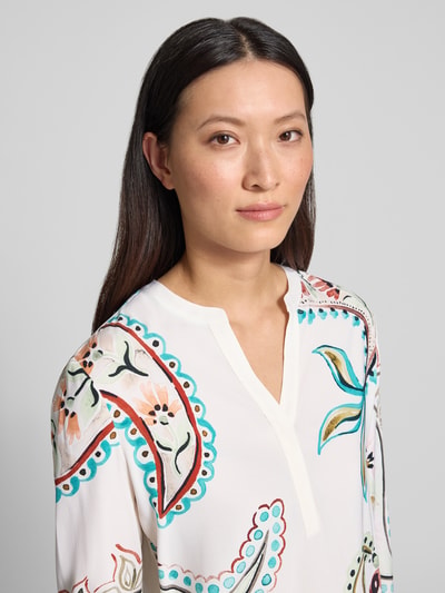 Frauenmodel trägt Relaxed Fit Tunika-Bluse mit Allover-Paisley-Print von HERZENS in Weiß mit bunten Details, Tunika-Ausschnitt, Langärmelig, Vorderseite, Nahaufnahme Oberkörper. Rückenlänge 70 cm, Ärmellänge 64 cm, verlängerte Rückseite.