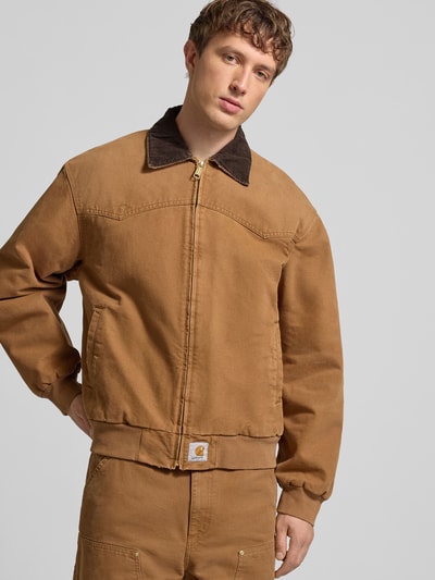 Męski model nosi Kurtkę z zamkiem błyskawicznym i detalem z logo od Carhartt Work In Progress z bawełny ekologicznej, krój regular fit, wykładany kołnierz, wpuszczane kieszenie, długość tyłu w rozmiarze M: 70 cm, szerokość ramion w rozmiarze M: 56 cm, szerokość klatki piersiowej w rozmiarze M: 65 cm, długość rękawów w rozmiarze M: 67 cm, brązowe kolory, widoczne faktury tkaniny, całkowity widok z przodu.