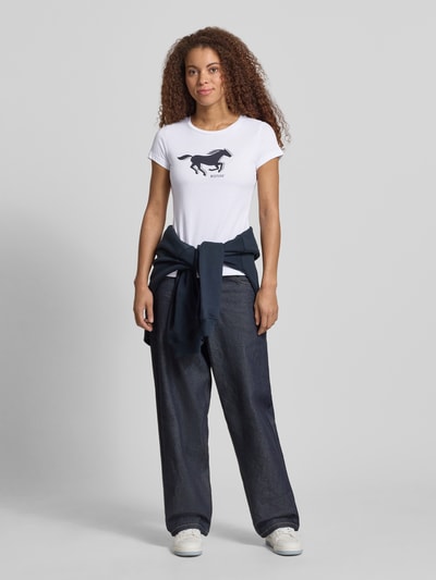 Modelka prezentuje damski t-shirt z bawełny Mustang, krój regular fit, model 'Loa' z nadrukiem z logo, okrągły dekolt, krótki rękaw, długość tyłu w rozmiarze S: 60 cm, widok całej sylwetki z przodu.