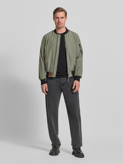 Männermodel trägt Bomberjacke mit Label-Patch und Eingrifftaschen von Original Bombers in Oliv, Gerader-Schnitt, Ganzkörperansicht von vorne, Reißverschluss, Reißverschlusstasche am Ärmel, Druckknopfverschluss, Gerippte-Abschlüsse, Rückenlänge 67 cm, Schulterbreite 43 cm, Brustweite 53 cm, Ärmellänge 68 cm bei Größe M.