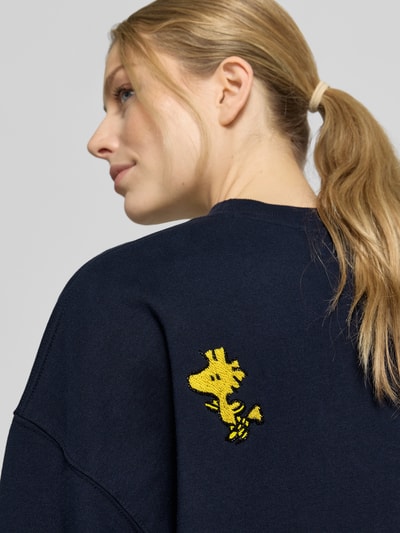 Frauenmodel trägt Loose Fit Sweatshirt aus reiner Baumwolle Modell 'JUMBRELLA EMBO' von Essentiel Antwerp in Dunkelblau mit gelbem Motiv-Print, Pailletten und Perlen, unifarbene Sweat-Optik, Nahaufnahme Oberkörper von schräg hinten.