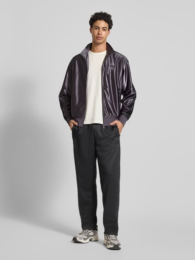 Männermodel trägt Sweatjacke mit Stehkragen von adidas Originals in Regular Fit, Reißverschluss, dunkler Glanzstoff, Ganzkörperansicht von vorne.