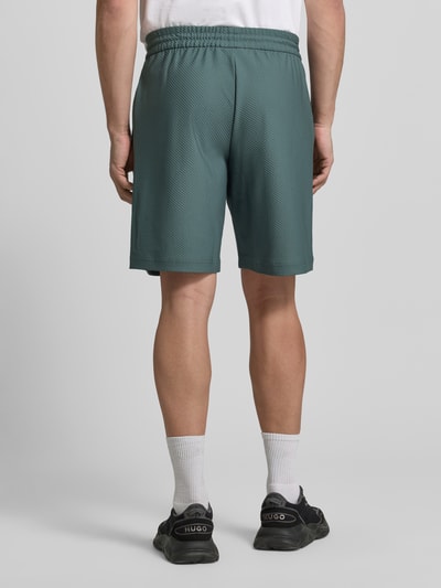 Mężczyzna nosi Regular Fit Shorts mit Strukturmuster od EA7 Emporio Armani, zielony kolor, elastyczny materiał, widok całej sylwetki z tyłu. Szerokość w rozmiarze M: 41 cm, długość nogawki od wewnątrz: 26 cm.